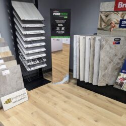 Mohawk PureTech flooring samples on display, with an information banner detailing the product's scratch resistance and PVC-free material, against a backdrop of a warmly lit flooring showroom.