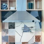 Colorful patchwork tile backsplash in a kitchen with a stainless steel hood and a pot filler faucet, featuring various patterned tiles and open shelves displaying pottery.